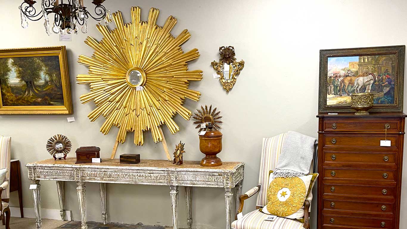 Inside Crown and Colony Antiques showroom showing a sunburst mirror over bleached console table and chair