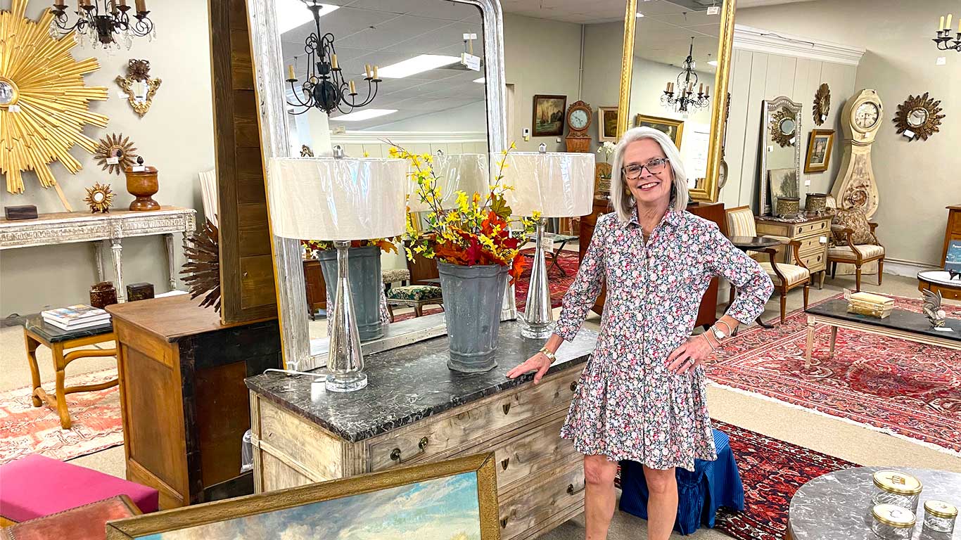 Inside Crown and Colony Antiques showroom with staff member standing in front of an antique buffet with lamps and mirror on top
